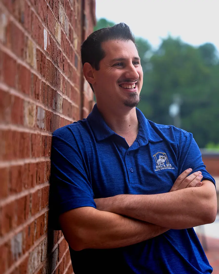George Karampoulis in his blue Owl’s Eye polo, arms folded, smiling beside a brick wall