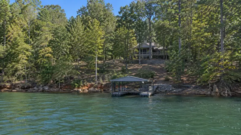 A lake house set back on a wooded slope, with a retaining wall and steps down to a covered boat dock, and red clay along the shoreline