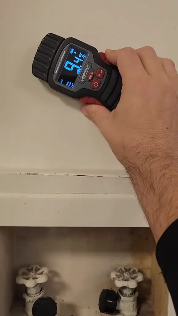 A moisture meter held against a wall above two plumbing shut-off valves, showing a reading