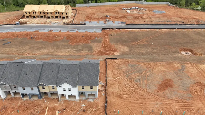 Aerial view of a new-home community under construction, with finished townhomes, framed buildings and red-clay lots